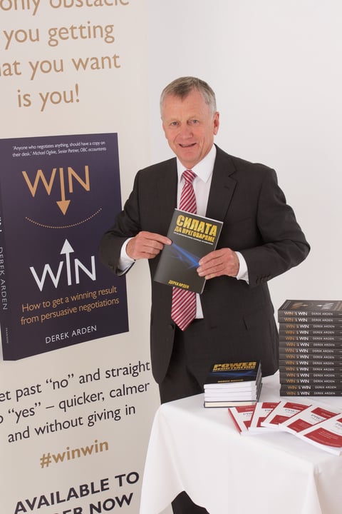 Man in dark suit and striped tie holding business book at table with stacked copies and promotional poster displaying WIN text
