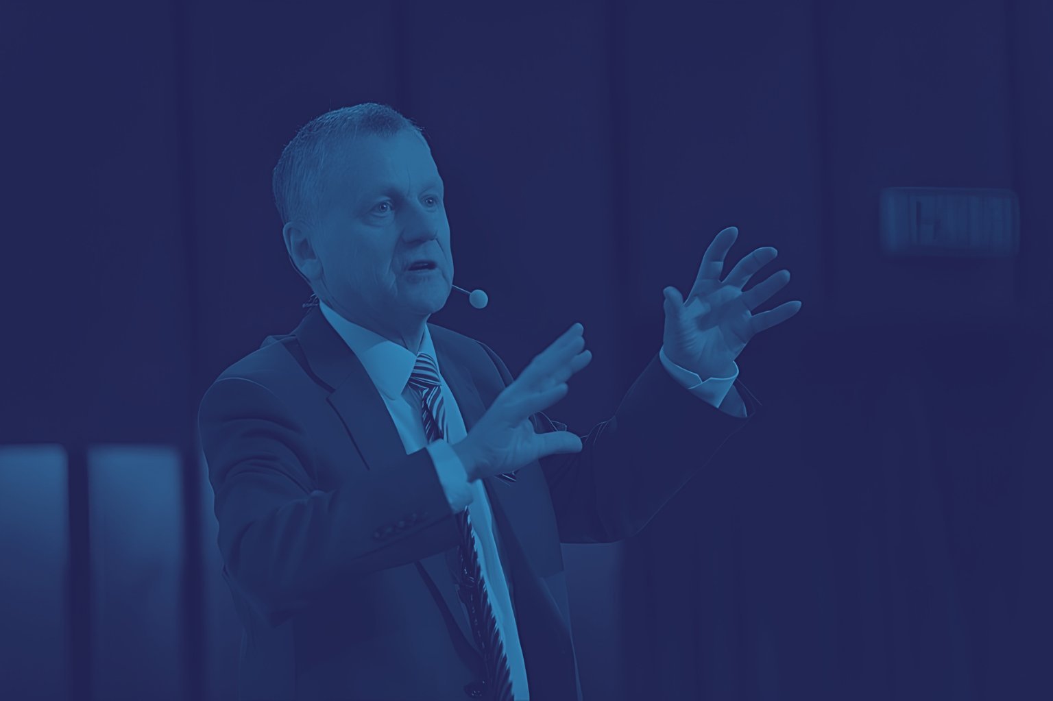 Man in business suit gesturing while speaking, toned in blue light against a dark background