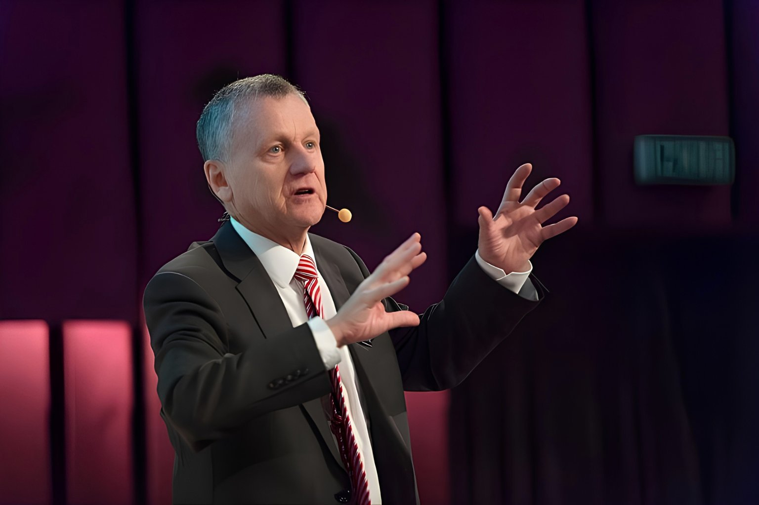 Middle-aged man in dark suit speaking passionately on stage with hands gesturing, wearing a headset microphone against a purple curtain backdrop